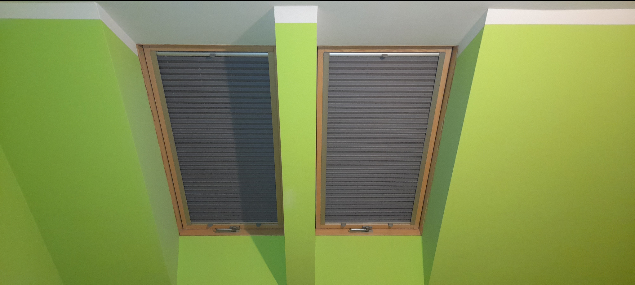 Dwa okna dachowe z szarymi plisami na tle zielonej ściany. Widok od dołu, podkreślający kąt nachylenia okien i dopasowanie plis do ich kształtu.