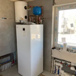 Biała pompa ciepła Nibe z zasobnikiem Galmet zamontowana w pomieszczeniu wykończonym płytkami, widoczne instalacje podłogowe i okno z parapetem z pianobetonu z narzędziami i rolką papieru.