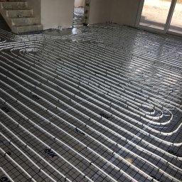 Kamil Więcław - Białe rury ogrzewania podłogowego ułożone na czarnej siatce w surowym wnętrzu domu w trakcie budowy, widoczne schody i okno balkonowe.