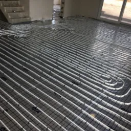 Białe rury ogrzewania podłogowego ułożone na czarnej siatce w surowym wnętrzu domu w trakcie budowy, widoczne schody i okno balkonowe.