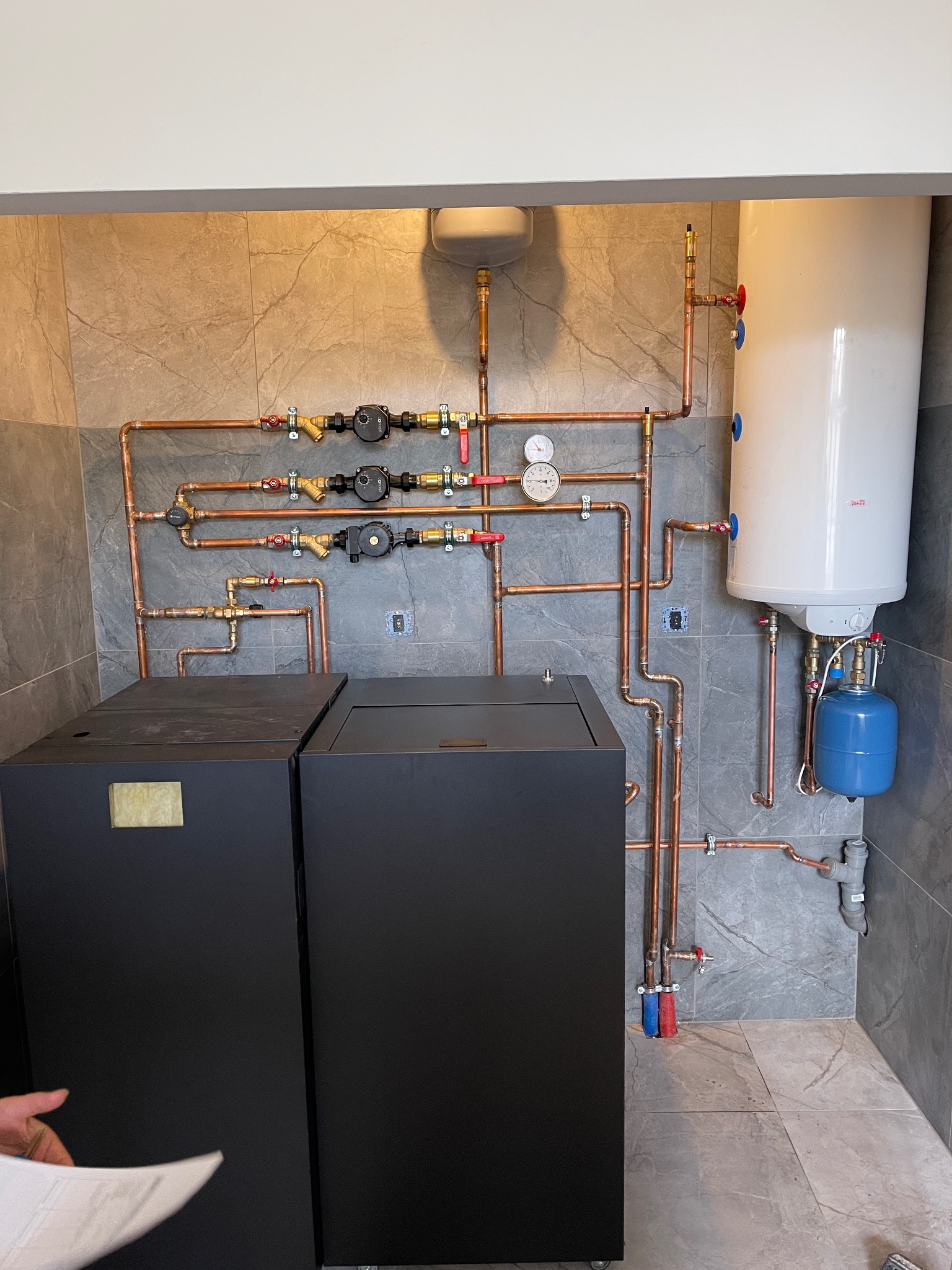 Kompletna instalacja grzewcza w kotłowni: dwa czarne kotły, biały zasobnik ciepłej wody, niebieskie naczynie przeponowe, miedziane rury z armaturą na tle szarych płytek imitujących marmur.