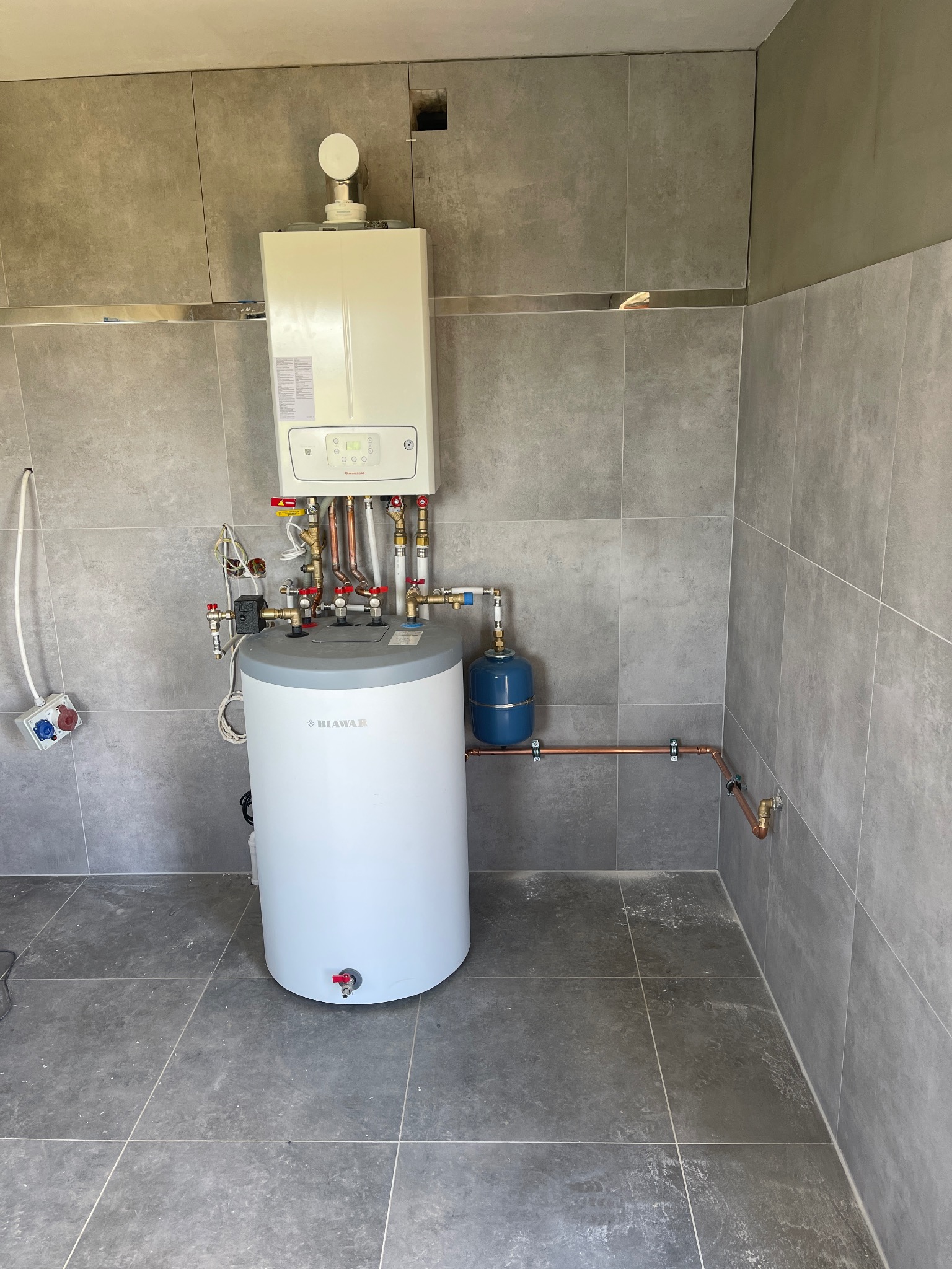 Biały piec gazowy z szarym zasobnikiem i niebieskim naczyniem wzbiorczym, podłączony miedzianymi rurami, zamontowany na tle szarych płytek w nowym pomieszczeniu.
