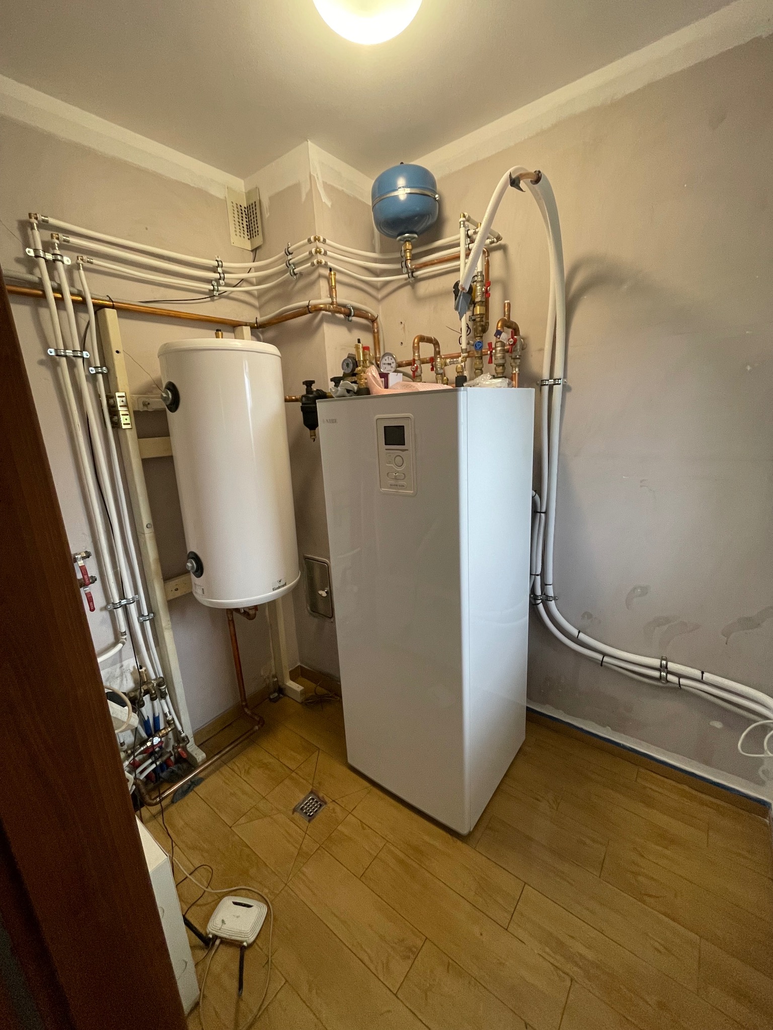 Biały piec grzewczy z podłączonymi rurami miedzianymi i plastikowymi w pomieszczeniu z drewnianą podłogą i szarymi ścianami.