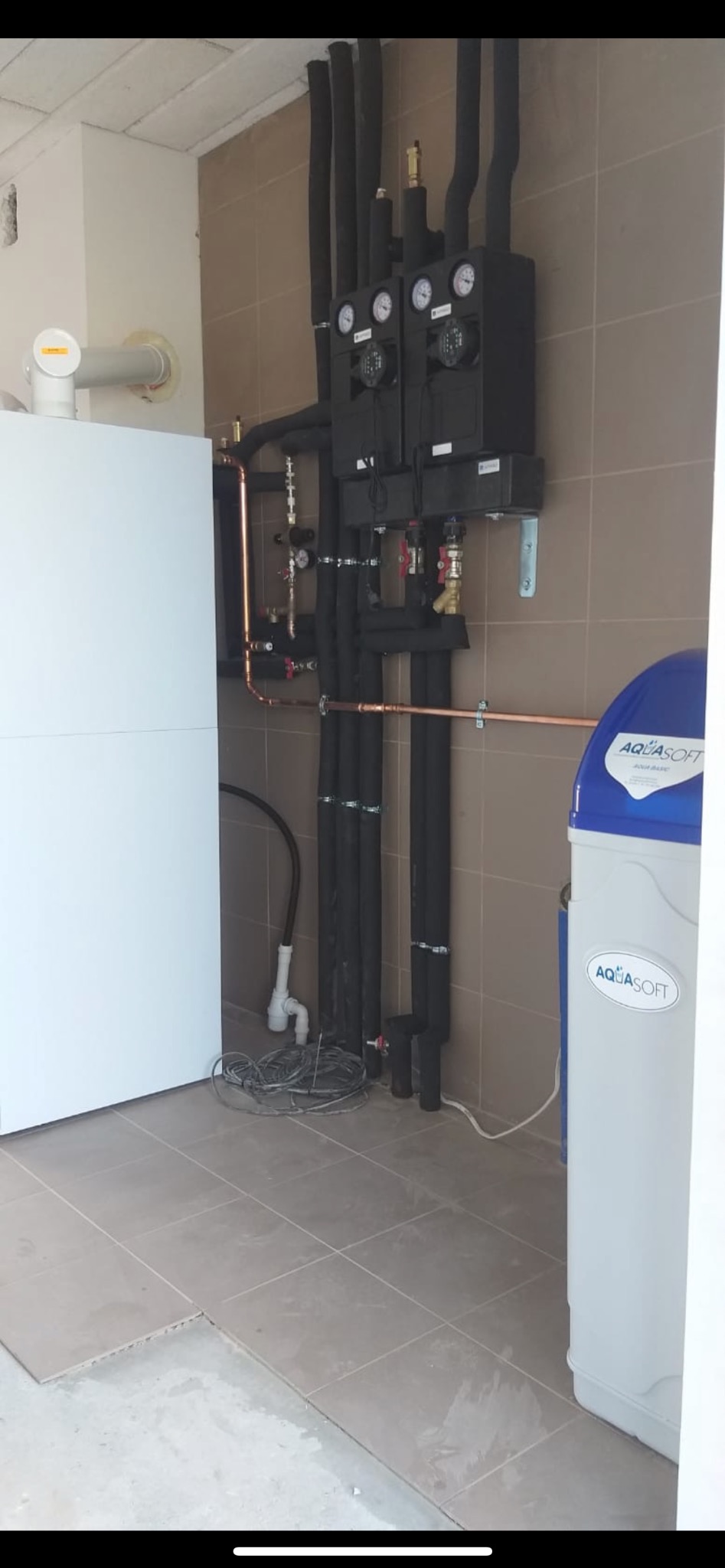 Kompletna instalacja grzewcza z czarnymi rurami izolacyjnymi, miedzianymi rurami i dwoma czarnymi modułami z manometrami zamontowanymi na ścianie z płytek, obok biały zbiornik Aquasoft i biały piec.