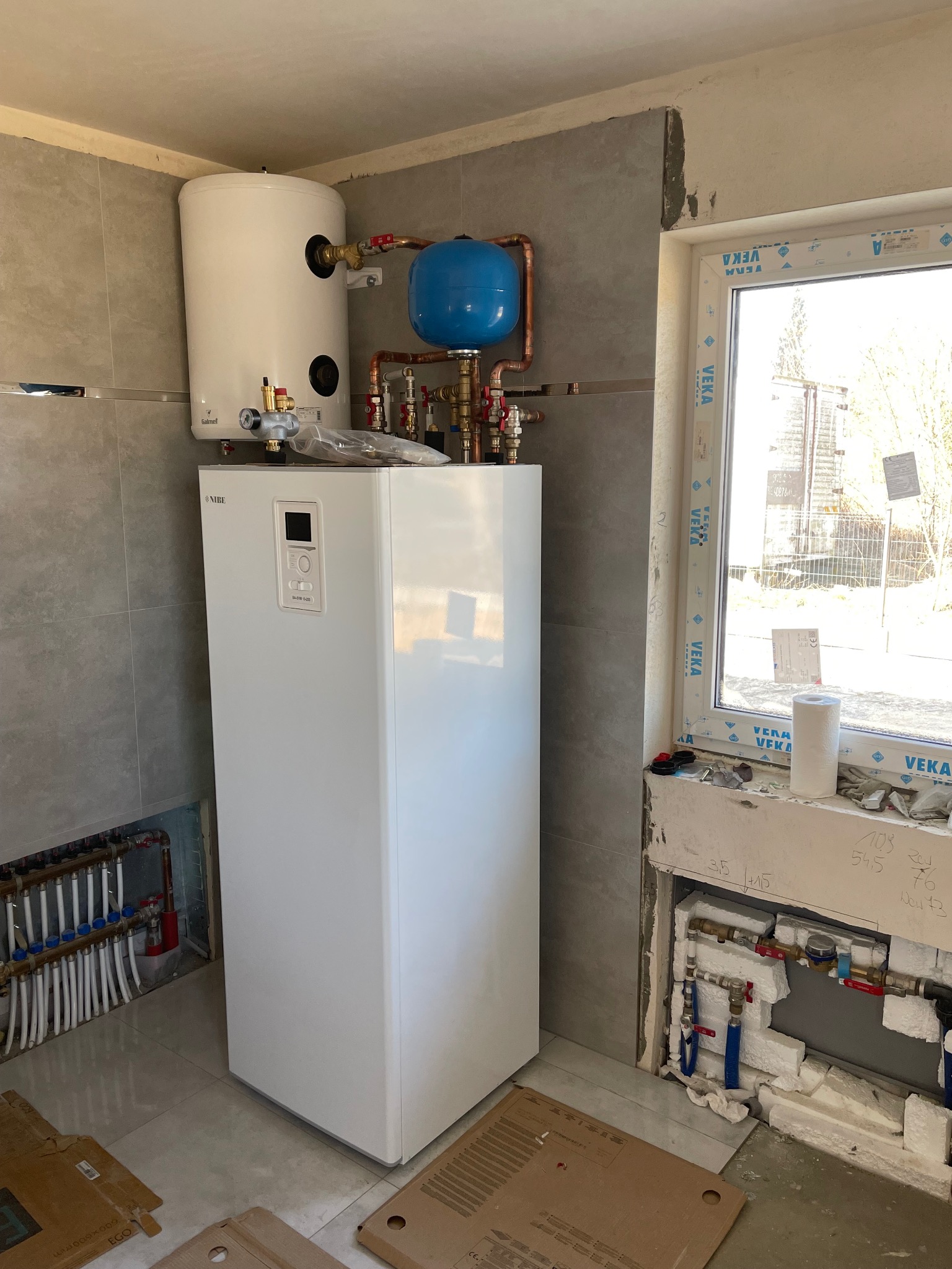 Biała pompa ciepła Nibe z zasobnikiem Galmet zamontowana w pomieszczeniu wykończonym płytkami, widoczne instalacje podłogowe i okno z parapetem z pianobetonu z narzędziami i rolką papieru.
