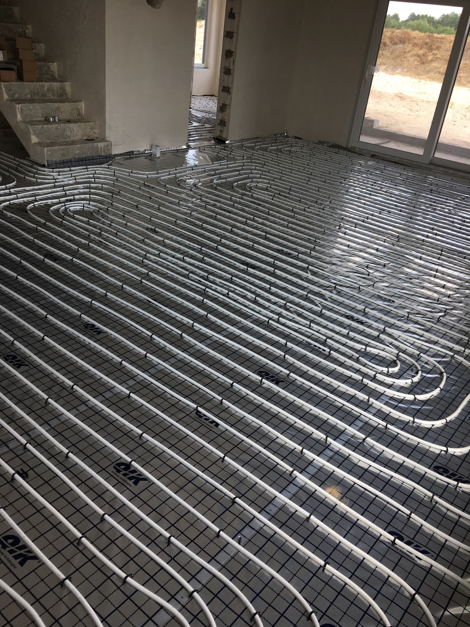 Białe rury ogrzewania podłogowego ułożone na czarnej siatce w surowym wnętrzu domu w trakcie budowy, widoczne schody i okno balkonowe.