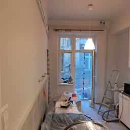 Wiktor Rosiński - Pokój w trakcie odświeżania: świeżo pomalowane ściany na biało, meble zabezpieczone folią malarską, widoczna drabina, wiadro z farbą i narzędzia malarskie, okno z widokiem na podwórze.