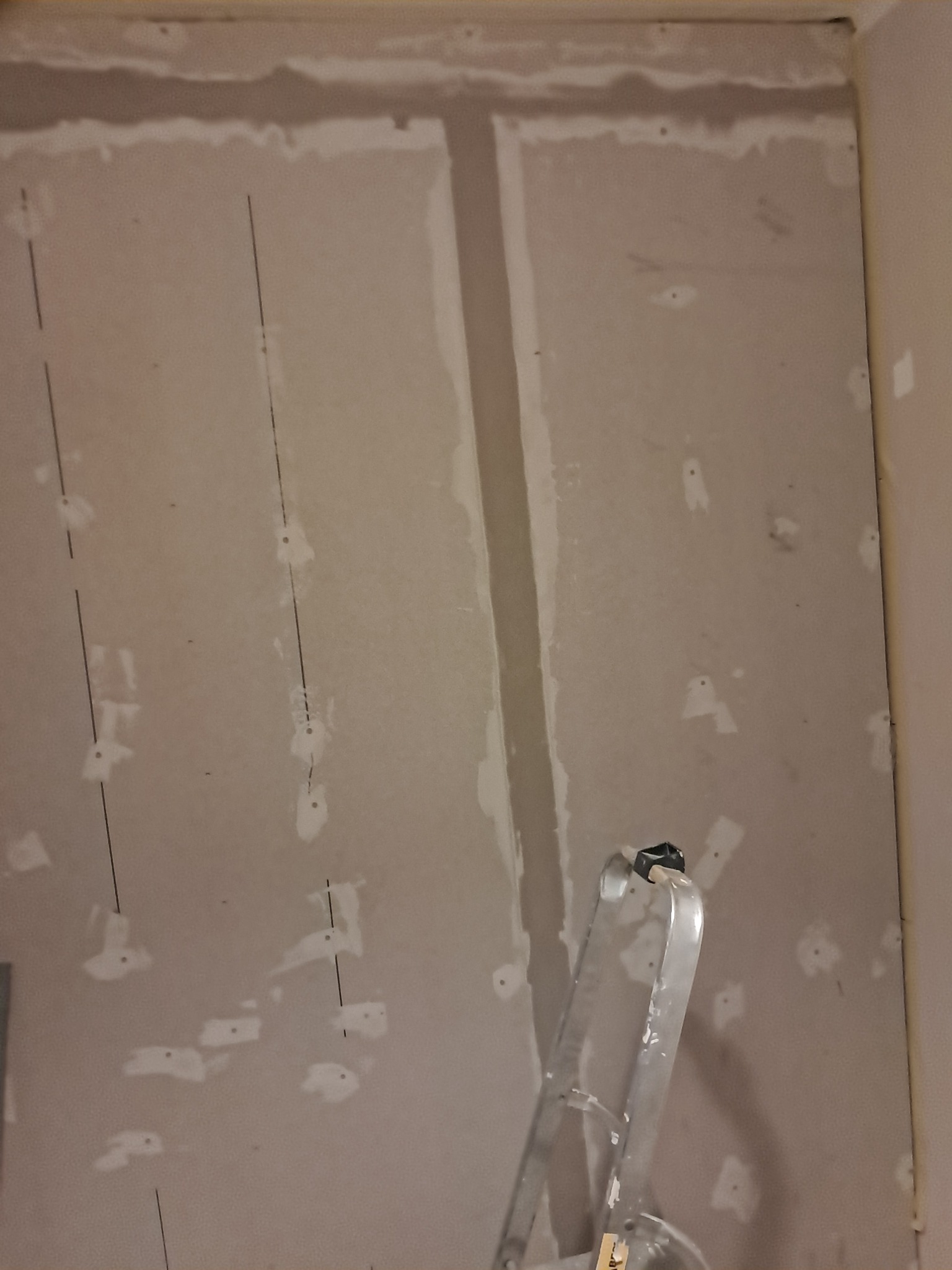 Ściana z płyt karton gips, widoczne szpachlowane łączenia i ślady po wkrętach, pionowe linie wyznaczające punkty mocowania, fragment drabiny aluminiowej w dolnym rogu.