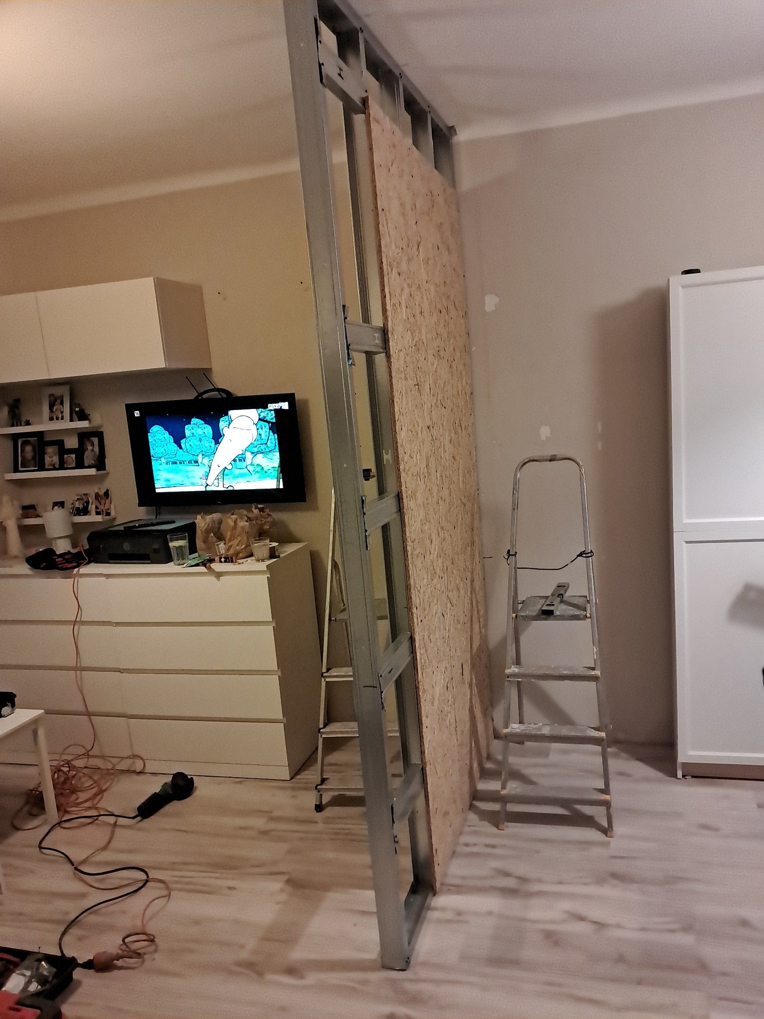 Montaż ścianki działowej z płyty OSB na metalowym stelażu w pokoju, widoczna drabina, narzędzia i meble w tle.