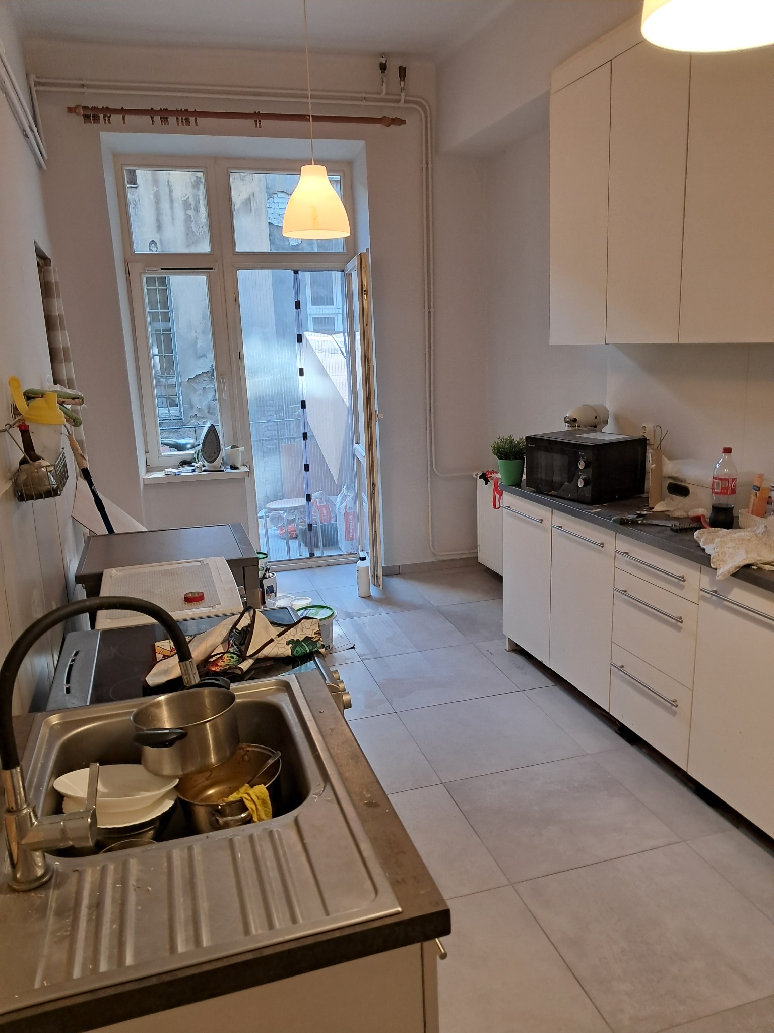 Wnętrze kuchni w trakcie remontu, widoczne niedokończone prace, brudny zlew pełen naczyń, białe szafki kuchenne i okno z widokiem na podwórko.