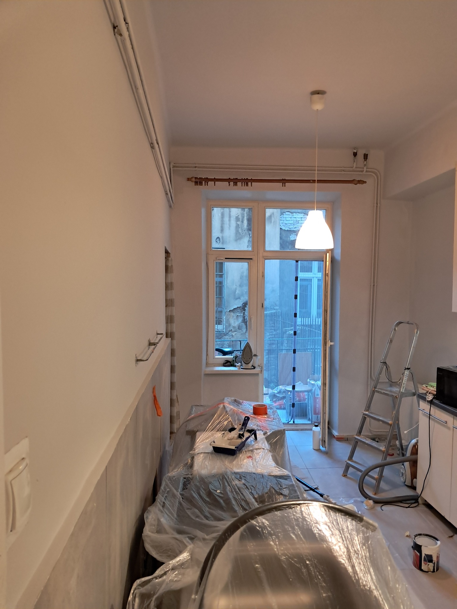 Pokój w trakcie odświeżania: świeżo pomalowane ściany na biało, meble zabezpieczone folią malarską, widoczna drabina, wiadro z farbą i narzędzia malarskie, okno z widokiem na podwórze.
