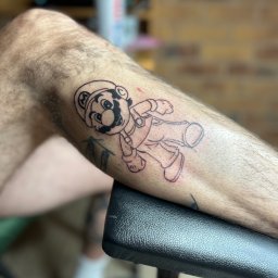 Blackheart - Świeżo wykonany, czarno-biały tatuaż na łydce przedstawiający postać Mario z gry Super Mario Bros., leżący na czarnym, skórzanym podłożu.