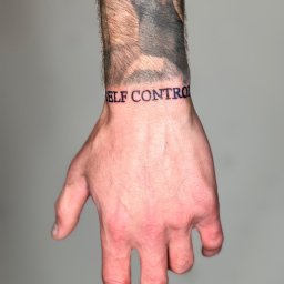 Blackheart - Męska dłoń z tatuażem 'SELF CONTROL' wokół nadgarstka i fragmentem innego tatuażu na przedramieniu, sfotografowana na neutralnym szarym tle.