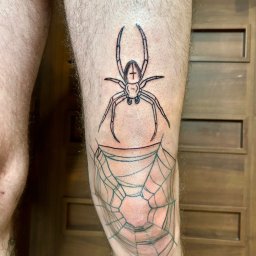 Blackheart - Czarno-biały tatuaż na nodze przedstawiający pająka z krzyżem na odwłoku nad pajęczyną, widoczny na tle drewnianych drzwi.