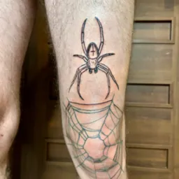 Czarno-biały tatuaż na nodze przedstawiający pająka z krzyżem na odwłoku nad pajęczyną, widoczny na tle drewnianych drzwi.