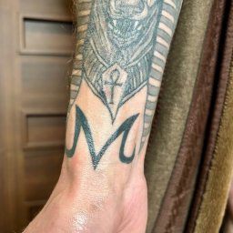 Blackheart - Szczegółowe ujęcie tatuażu na przedramieniu przedstawiającego egipski motyw z symbolem ankh i literą M, wykonane w czarnym tuszu.