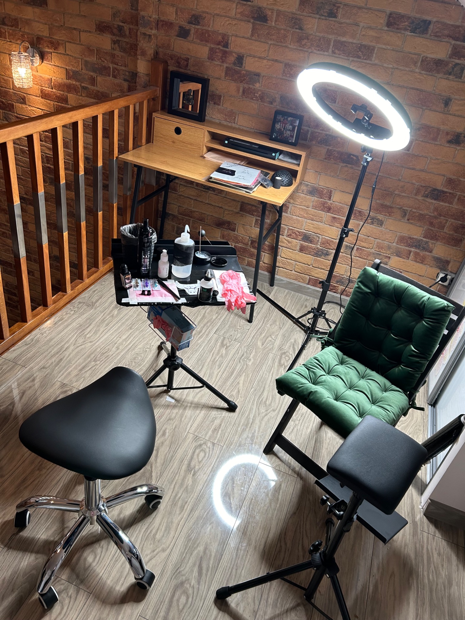 Stanowisko pracy stylisty z oświetleniem ring light, biurkiem, taboretem siodłowym i krzesłem z zieloną poduszką na tle ściany z cegły.