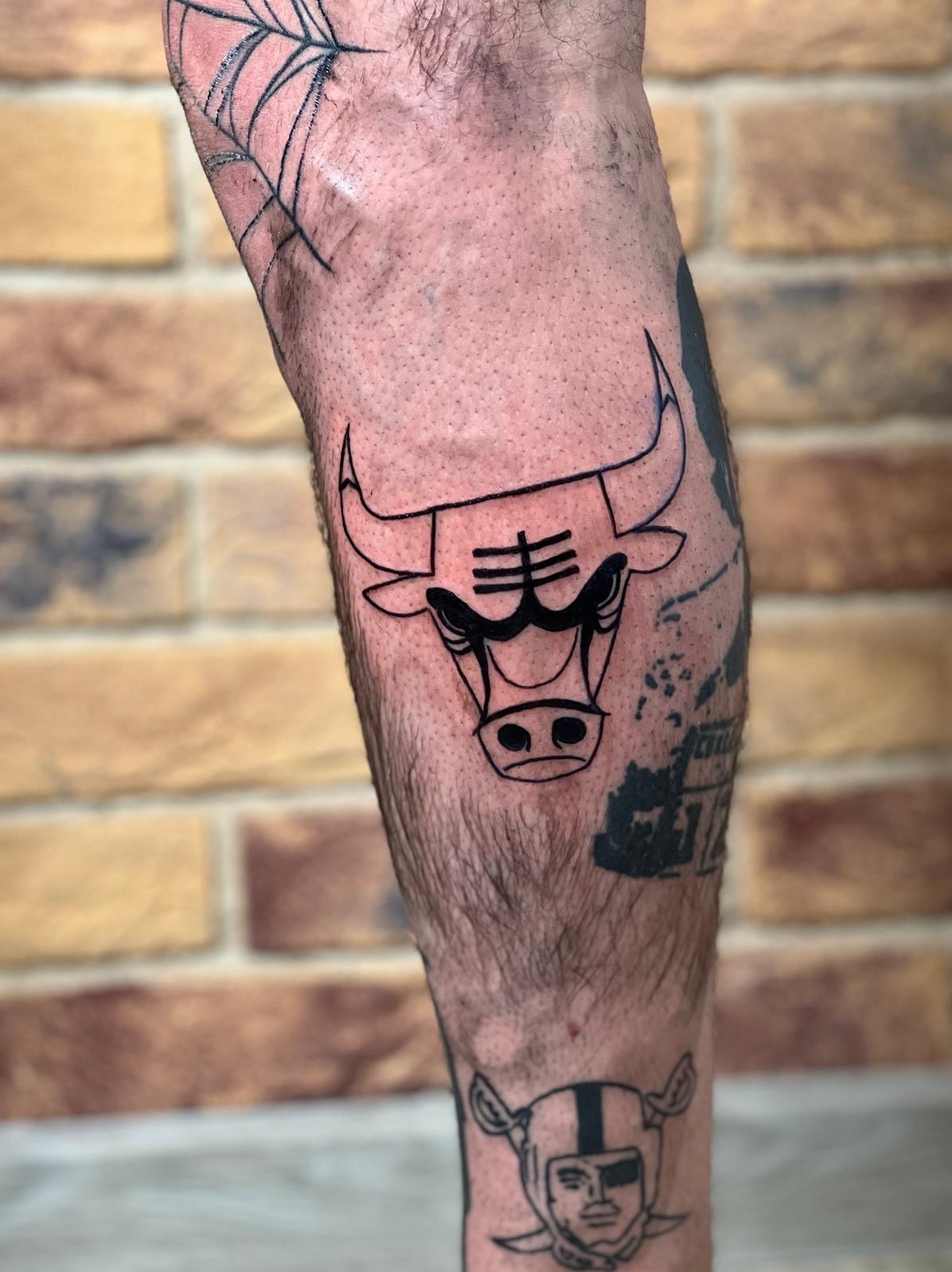 Tatuaż czarnym tuszem przedstawiający głowę byka na łydce mężczyzny, widoczny również tatuaż pajęczyny na kolanie i tatuaż głowy w hełmie na dole łydki, tło z cegieł.