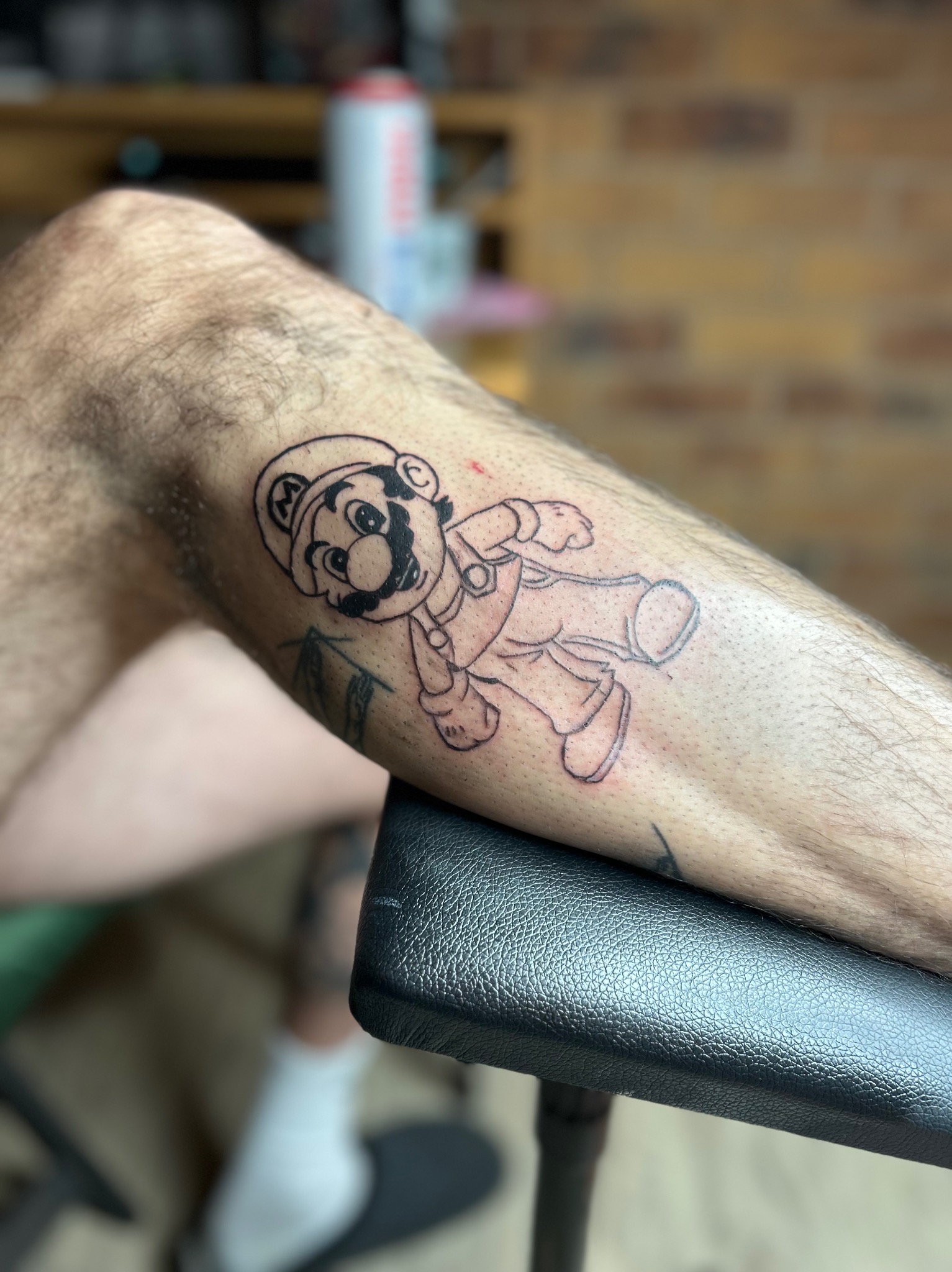 Świeżo wykonany, czarno-biały tatuaż na łydce przedstawiający postać Mario z gry Super Mario Bros., leżący na czarnym, skórzanym podłożu.