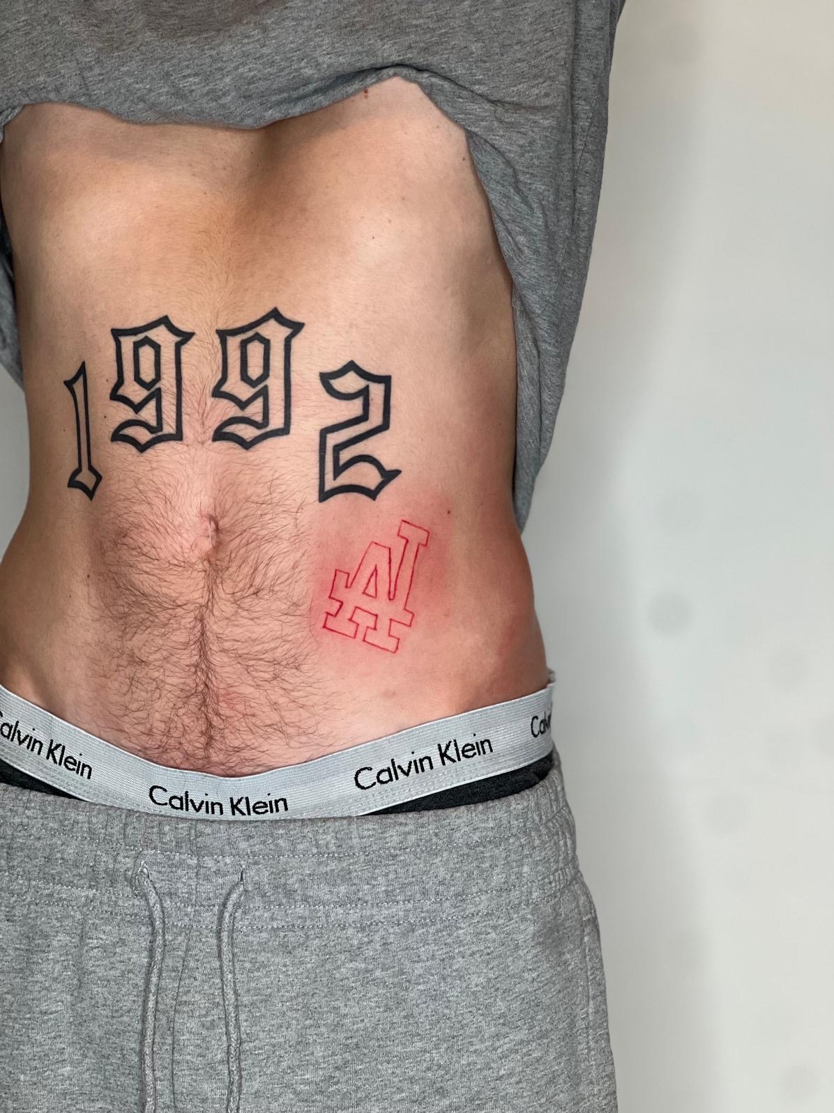 Męski tors z tatuażami: czarne cyfry 1992 nad pępkiem i czerwony kontur LA po prawej stronie, widoczna elastyczna guma spodni marki Calvin Klein.
