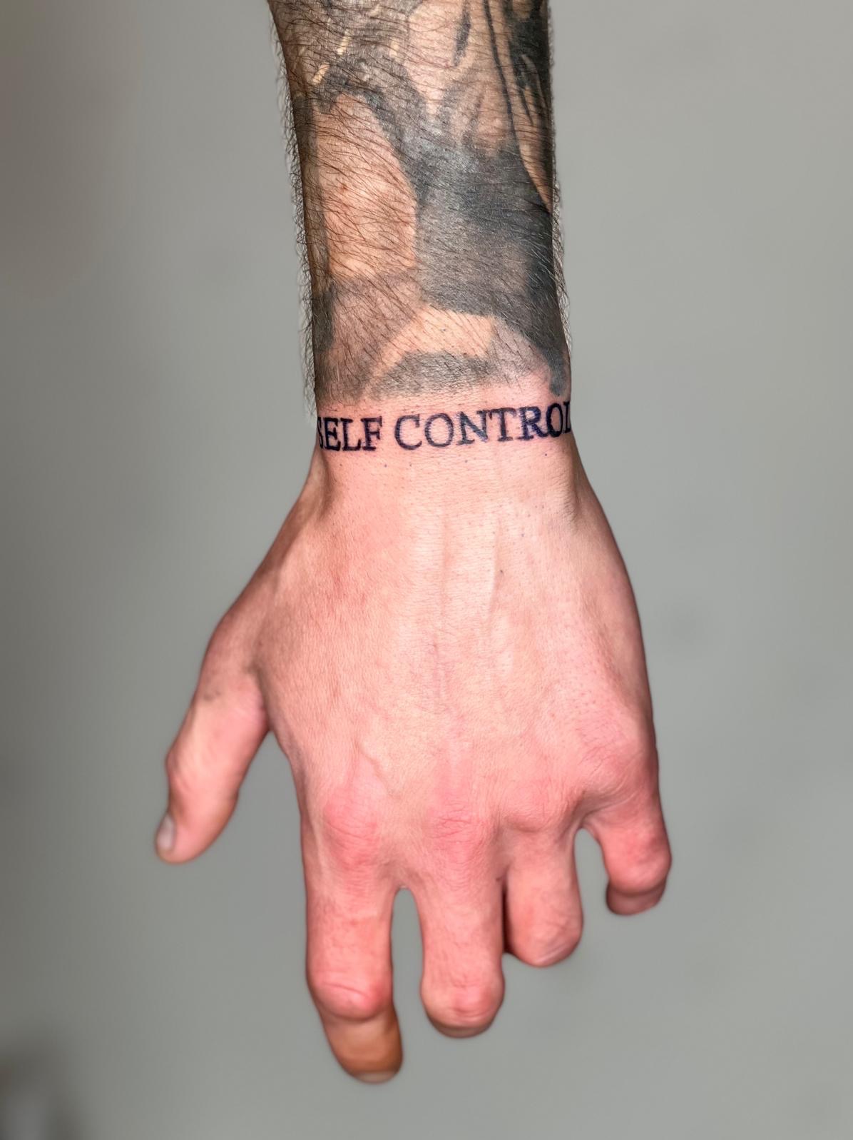 Męska dłoń z tatuażem 'SELF CONTROL' wokół nadgarstka i fragmentem innego tatuażu na przedramieniu, sfotografowana na neutralnym szarym tle.
