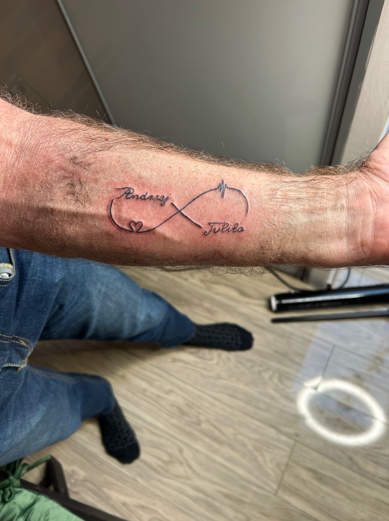 Tatuaż na przedramieniu przedstawiający symbol nieskończoności z imionami 'Andrzej' i 'Julita', połączony symbolem serca i linią EKG.