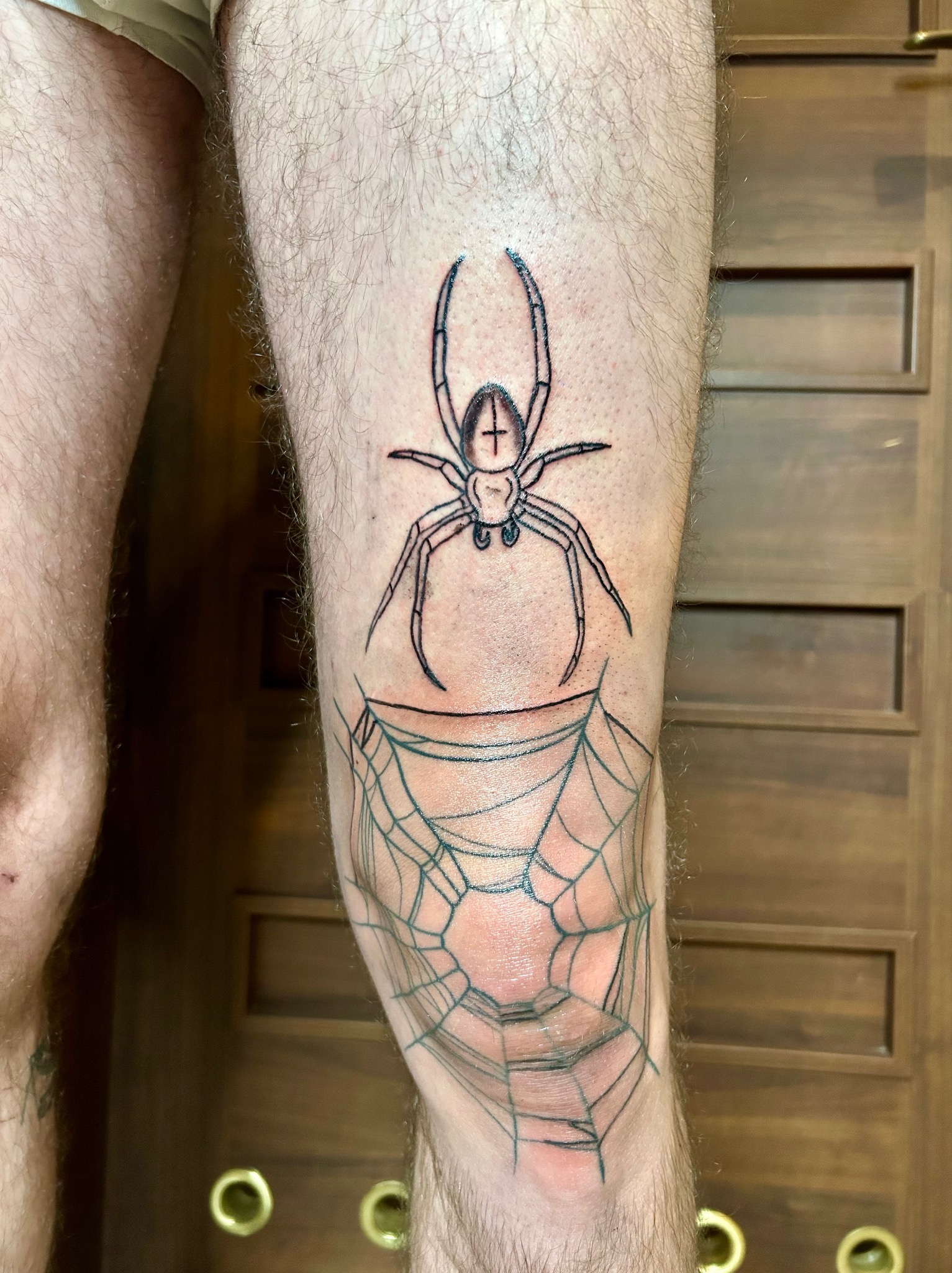 Czarno-biały tatuaż na nodze przedstawiający pająka z krzyżem na odwłoku nad pajęczyną, widoczny na tle drewnianych drzwi.