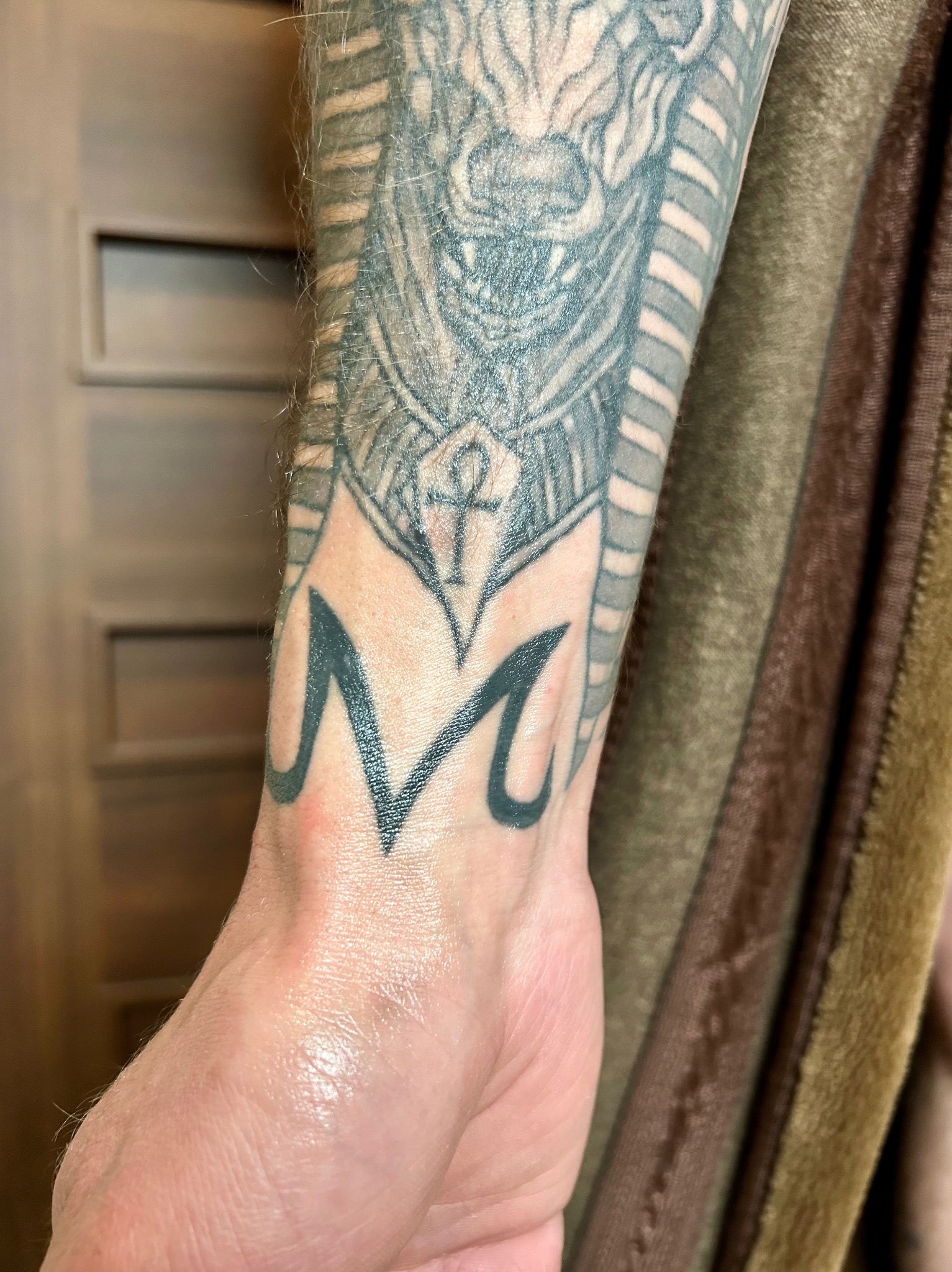 Szczegółowe ujęcie tatuażu na przedramieniu przedstawiającego egipski motyw z symbolem ankh i literą M, wykonane w czarnym tuszu.