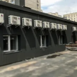 Rząd klimatyzatorów marki Gree zamontowanych na ścianie budynku biurowego z szarej blachy trapezowej, widok z zewnątrz, w tle budynek mieszkalny i fragment nieba.