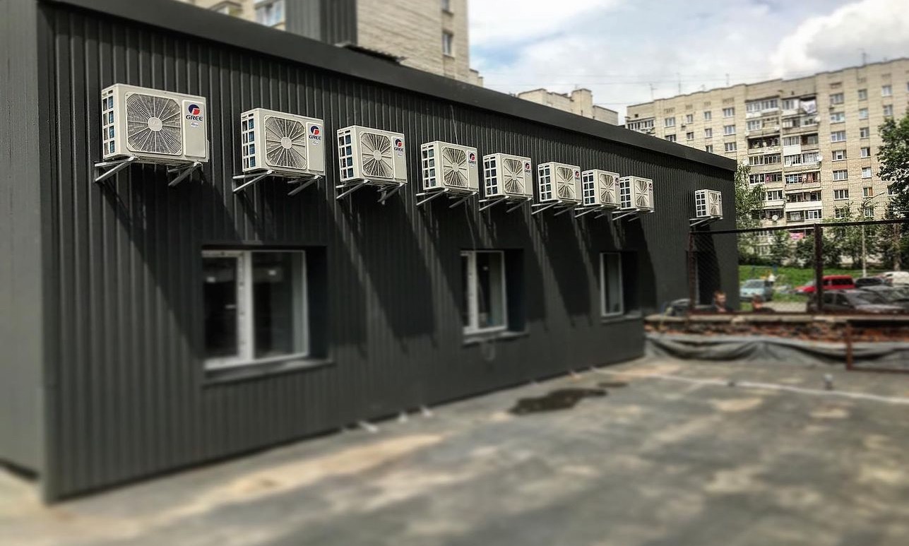 Rząd klimatyzatorów marki Gree zamontowanych na ścianie budynku biurowego z szarej blachy trapezowej, widok z zewnątrz, w tle budynek mieszkalny i fragment nieba.