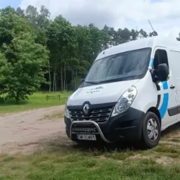 Biały Renault Master z niebieskimi akcentami i logotypem firmy na tle leśnej polany, z dodatkowym oświetleniem zamontowanym na przednim orurowaniu.