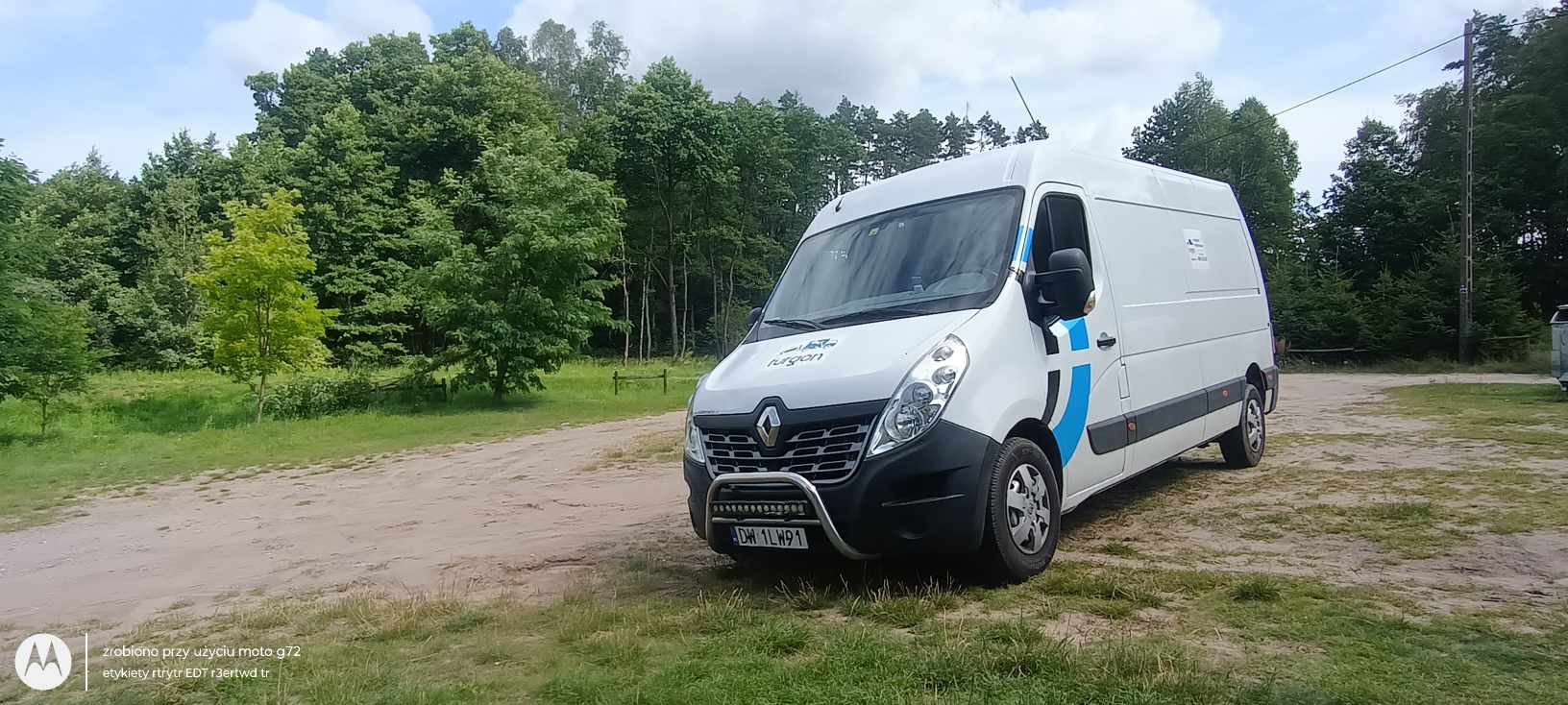 Biały Renault Master z niebieskimi akcentami i logotypem firmy na tle leśnej polany, z dodatkowym oświetleniem zamontowanym na przednim orurowaniu.