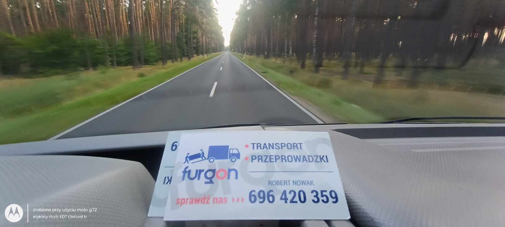 Widok zza kierownicy pojazdu na drogę prowadzącą przez las, z wizytówką firmy transportowej 'Furgon' na desce rozdzielczej, oferującej usługi transportowe i przeprowadzkowe. Numer telefonu: 696 420...