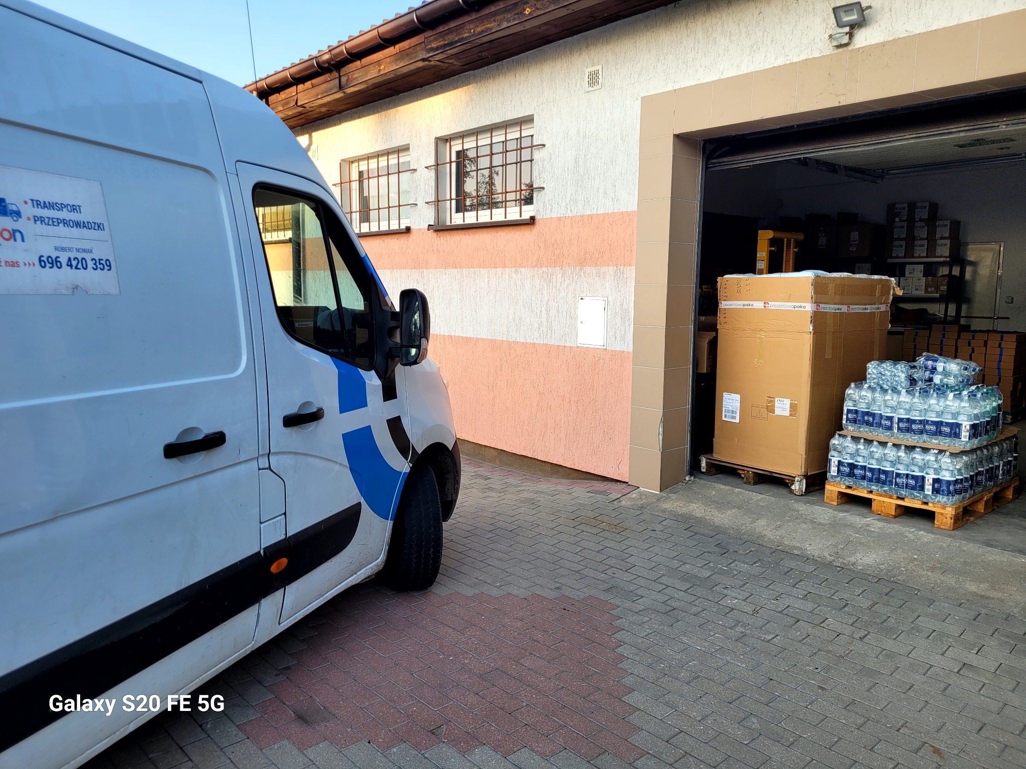 Biały samochód dostawczy z niebieskim logo firmy transportowej, zaparkowany przed otwartym garażem, w którym widoczne są kartony i palety z wodą mineralną.