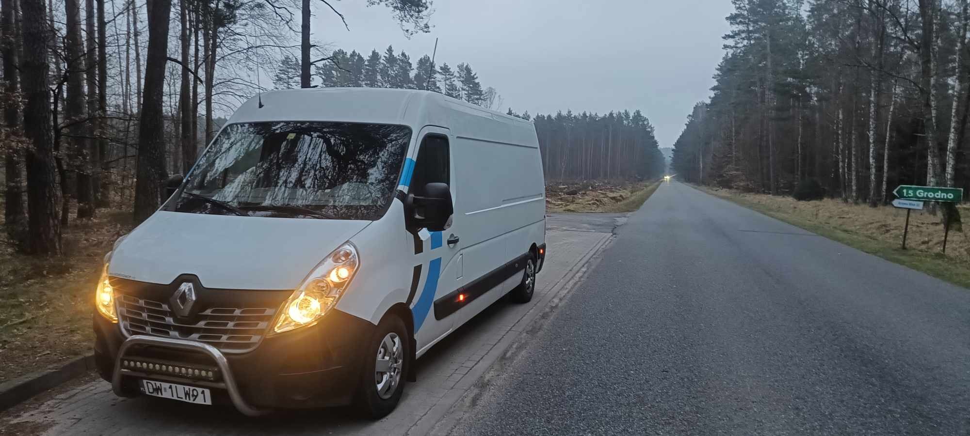 Biały bus Renault Master z niebieskimi akcentami parkuje na poboczu drogi w lesie; widoczny znak drogowy wskazujący kierunek na Grodno.