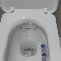 Iryna Zinchenko - Otwarta, biała toaleta z zawieszką zapachową w formie kolorowych kulek na krawędzi muszli.