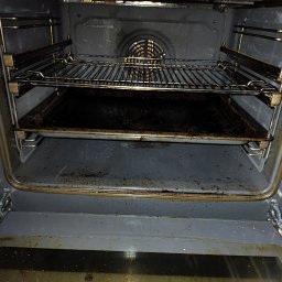 Iryna Zinchenko - Wnętrze brudnego piekarnika z rusztem i blachą, widoczne przypalone resztki jedzenia na ściankach i dnie.