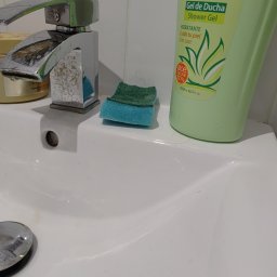 Iryna Zinchenko - Umywalka z chromowaną baterią pokrytą osadem, obok butelka żelu pod prysznic Aloe Vera i wilgotna, dwukolorowa gąbka do mycia.