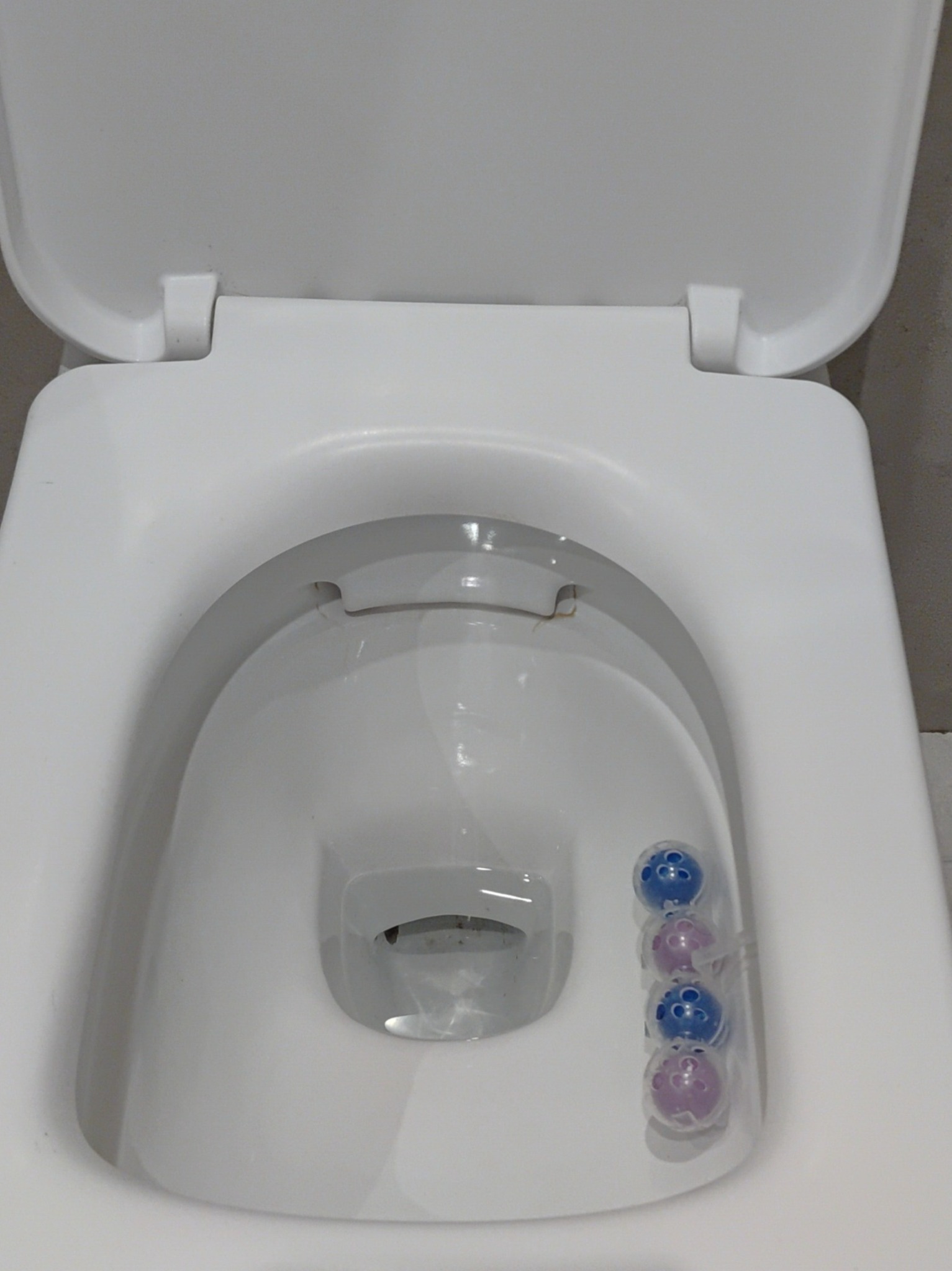 Otwarta, biała toaleta z zawieszką zapachową w formie kolorowych kulek na krawędzi muszli.