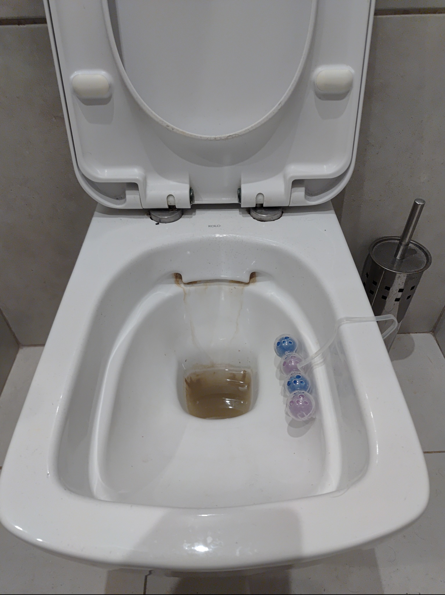 Brudna toaleta z otwartą klapą, widoczne osady i zawieszka odświeżająca z kolorowymi kulkami oraz szczotka do WC w metalowym pojemniku.