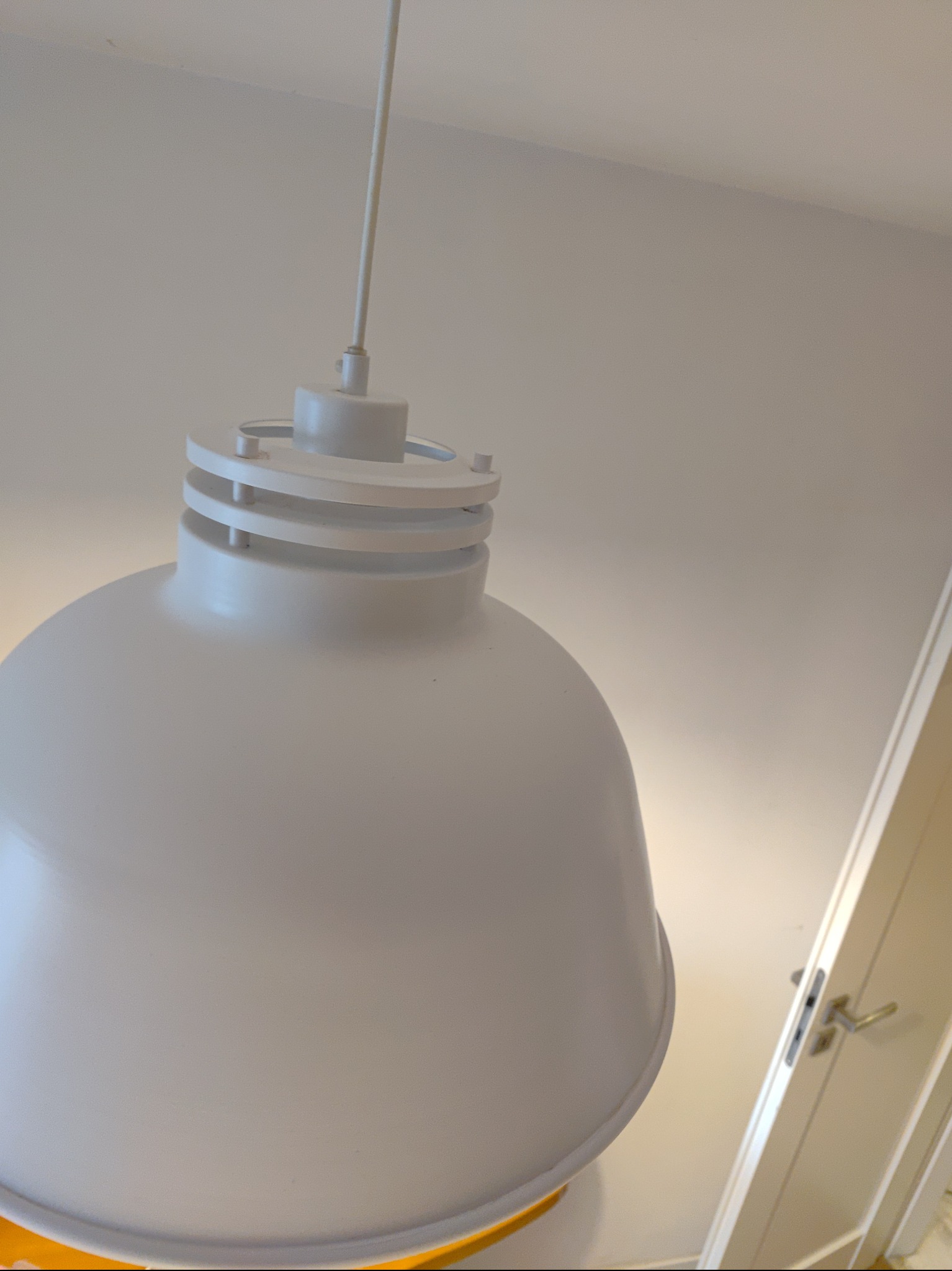 Duża, biała lampa wisząca z trzema pierścieniami na górze, zawieszona na białym kablu, widoczna na tle białego sufitu i fragmentu białych drzwi z klamką.