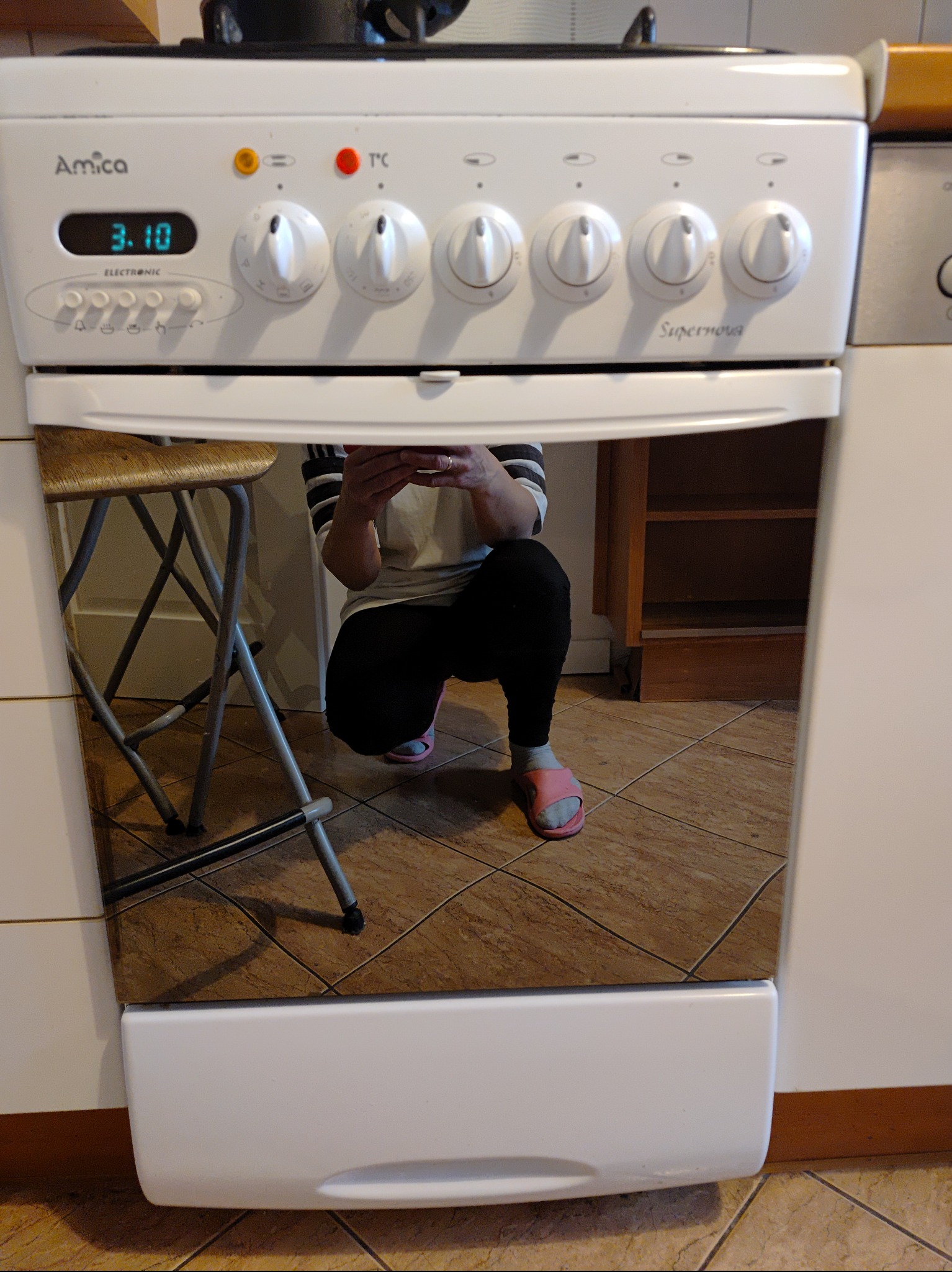 Błyszczące, odbijające się drzwiczki piekarnika Amica Supernova z widocznym odbiciem osoby robiącej zdjęcie i fragmentem krzesła.