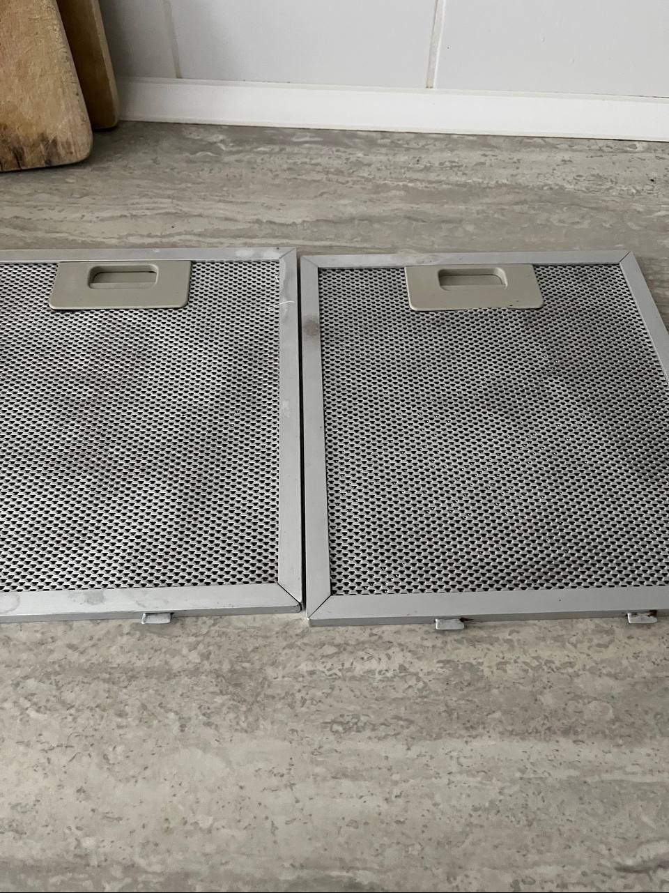 Dwa wyjęte, metalowe filtry okapu kuchennego z uchwytami, leżące na szarej, marmurkowej podłodze w pobliżu białej ściany.