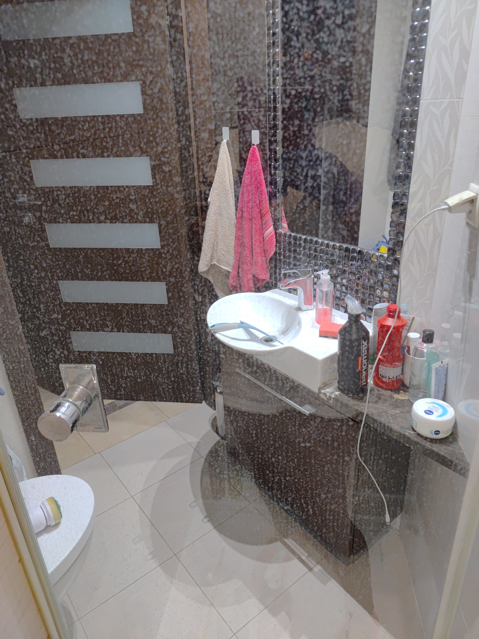 Zabrudzona kabina prysznicowa z widocznymi smugami i osadami na szklanych powierzchniach, obok umywalka z różnymi środkami czystości i gąbkami.