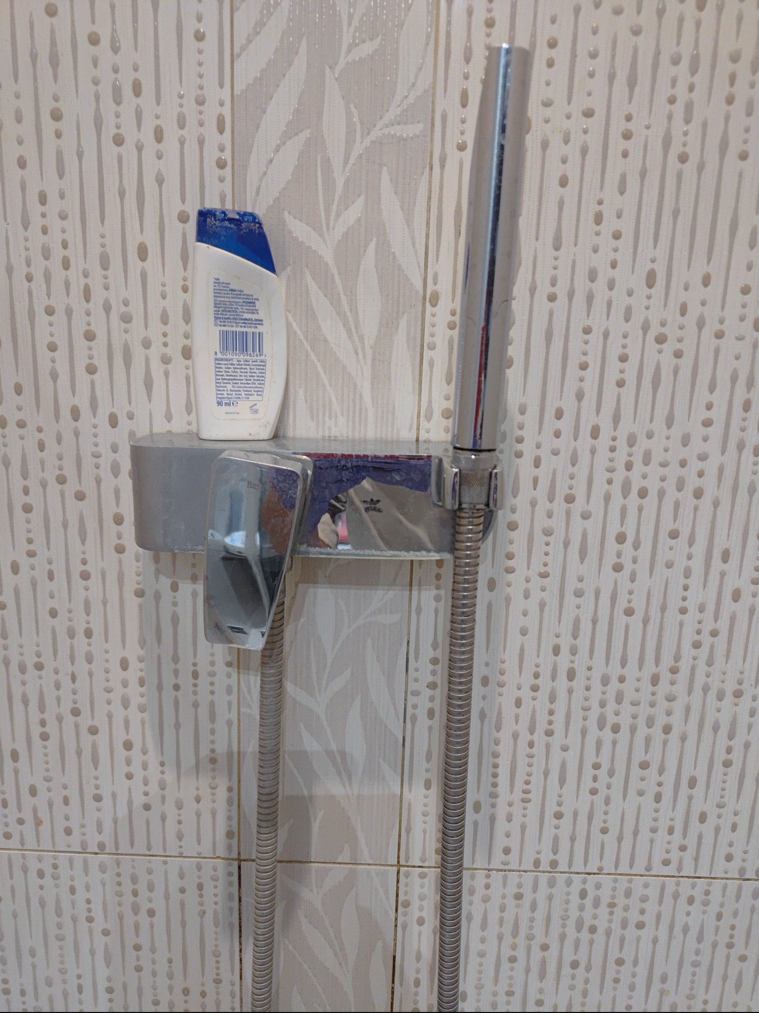 Półka prysznicowa z chromowaną słuchawką, wężem i butelką szamponu na tle beżowych płytek z wzorem kropli i liści.