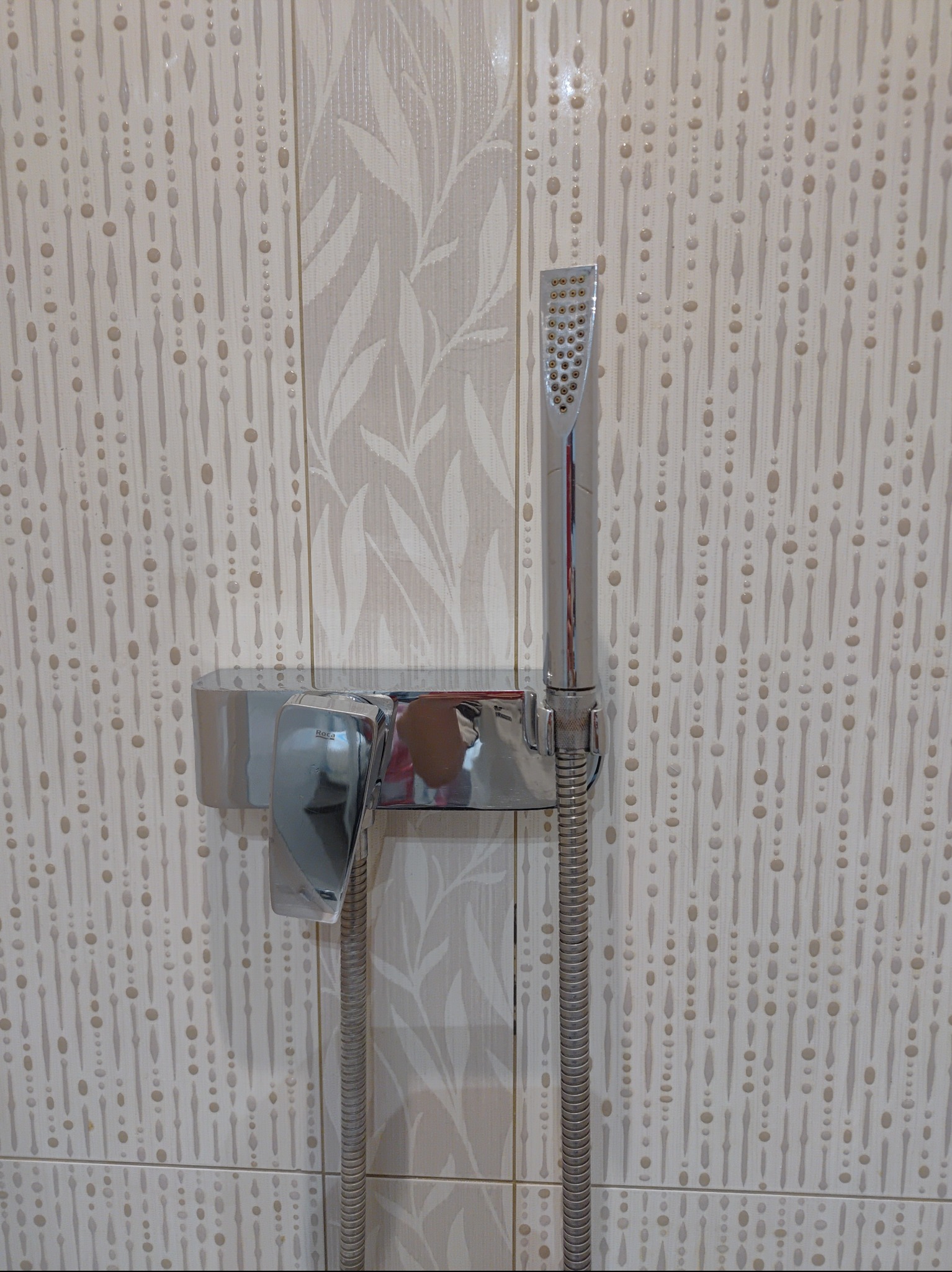 Chromowana bateria prysznicowa Roca zamontowana na beżowych płytkach z wzorem kropli i liści.