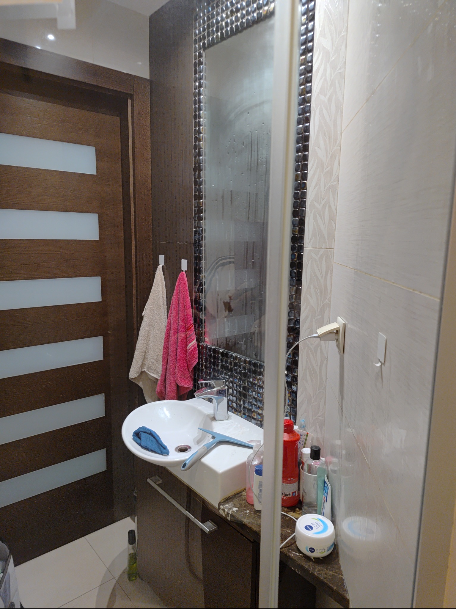 Umywalka z niebieską ściereczką i ściągaczką do wody obok, ręczniki na wieszakach, kosmetyki na blacie, widoczne ślady po myciu na lustrze.