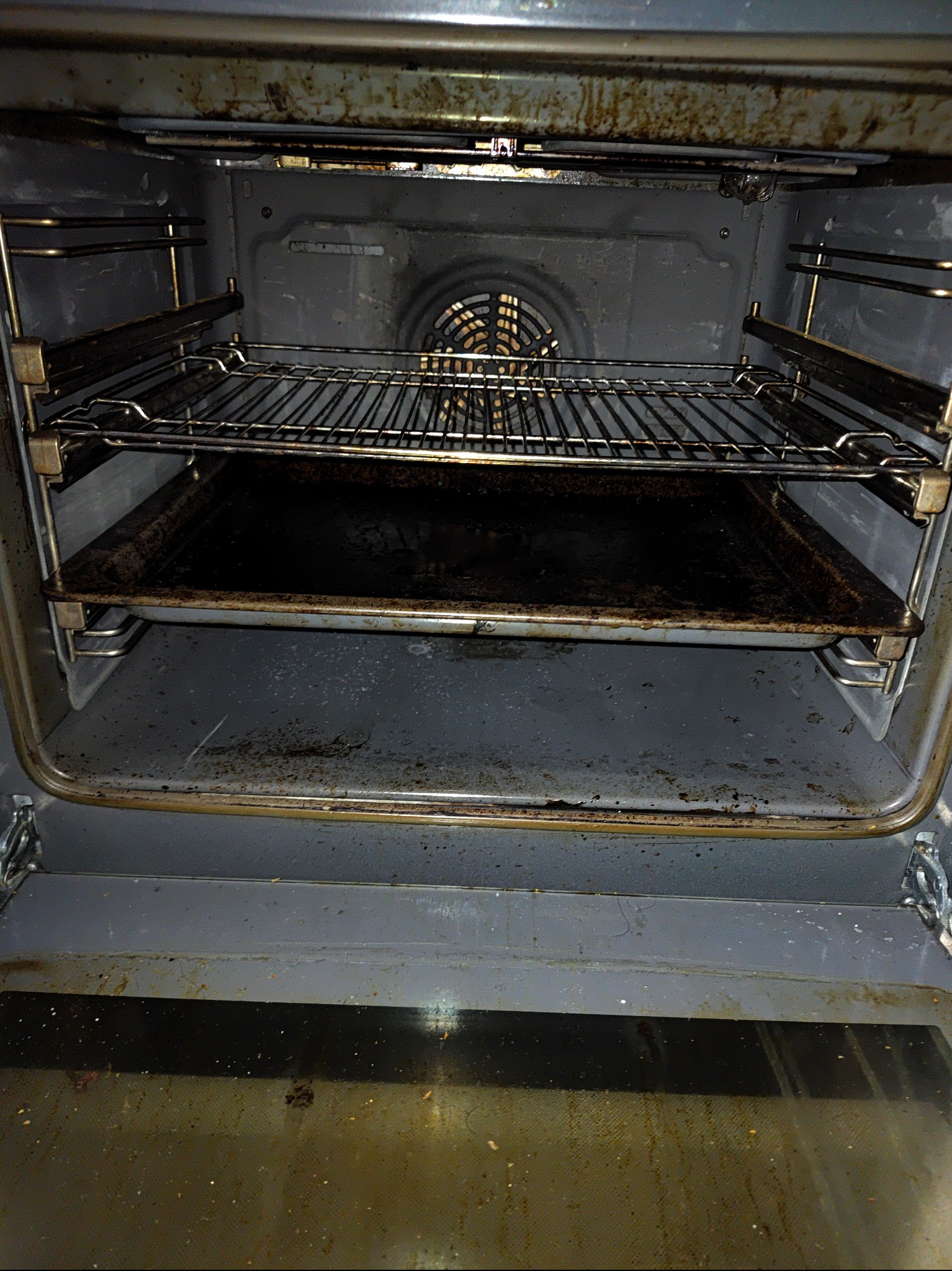 Wnętrze brudnego piekarnika z rusztem i blachą, widoczne przypalone resztki jedzenia na ściankach i dnie.
