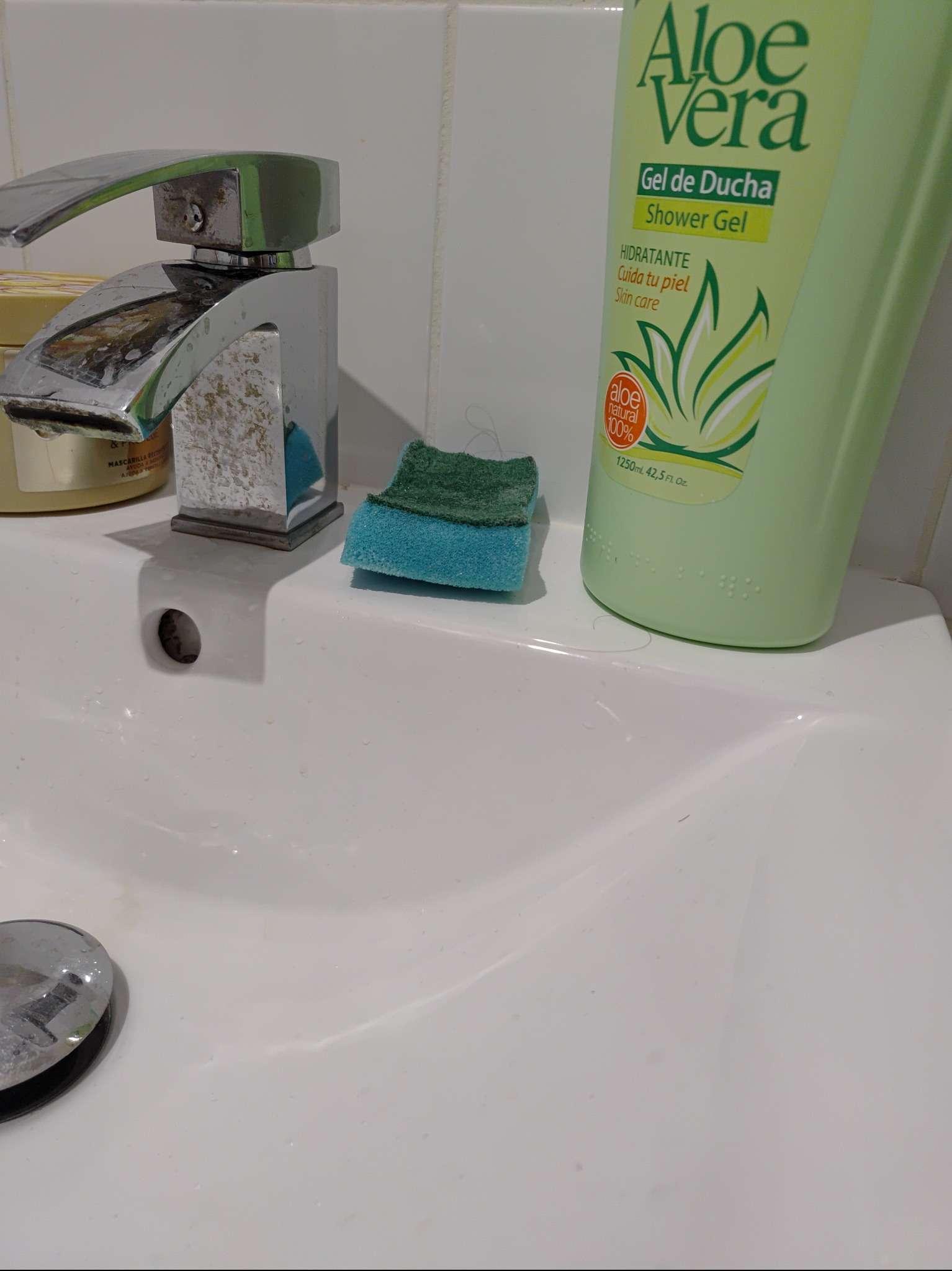 Umywalka z chromowaną baterią pokrytą osadem, obok butelka żelu pod prysznic Aloe Vera i wilgotna, dwukolorowa gąbka do mycia.