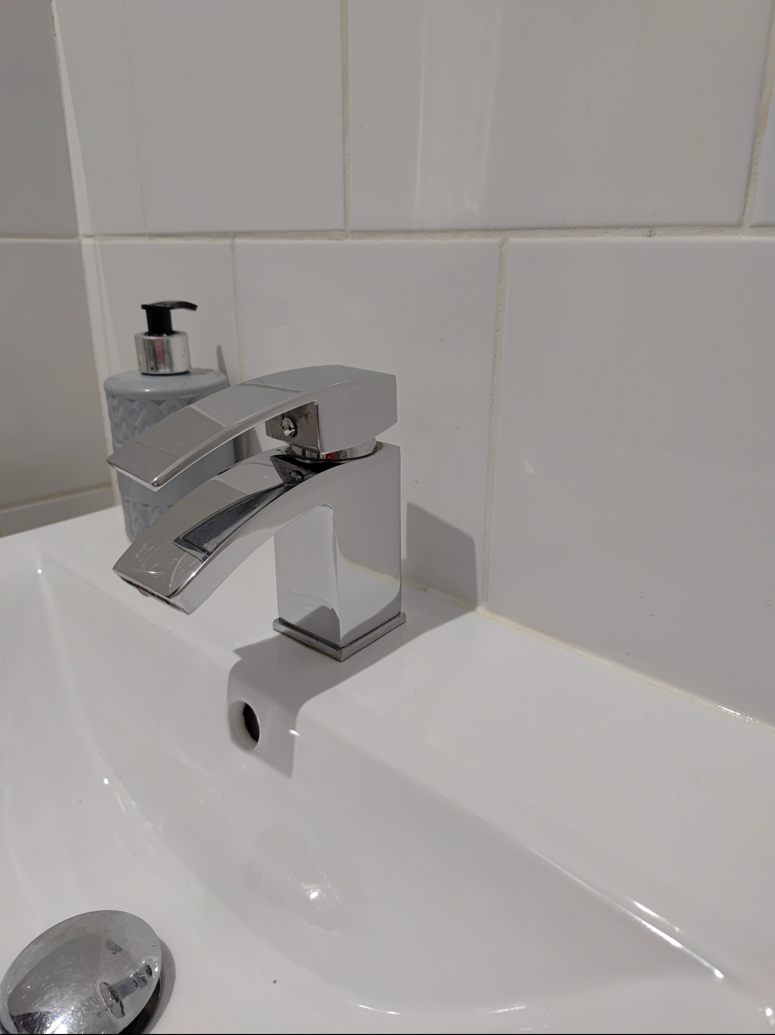 Błyszcząca, chromowana bateria umywalkowa o kwadratowym designie, obok szarego dozownika mydła w łazience z białymi płytkami.